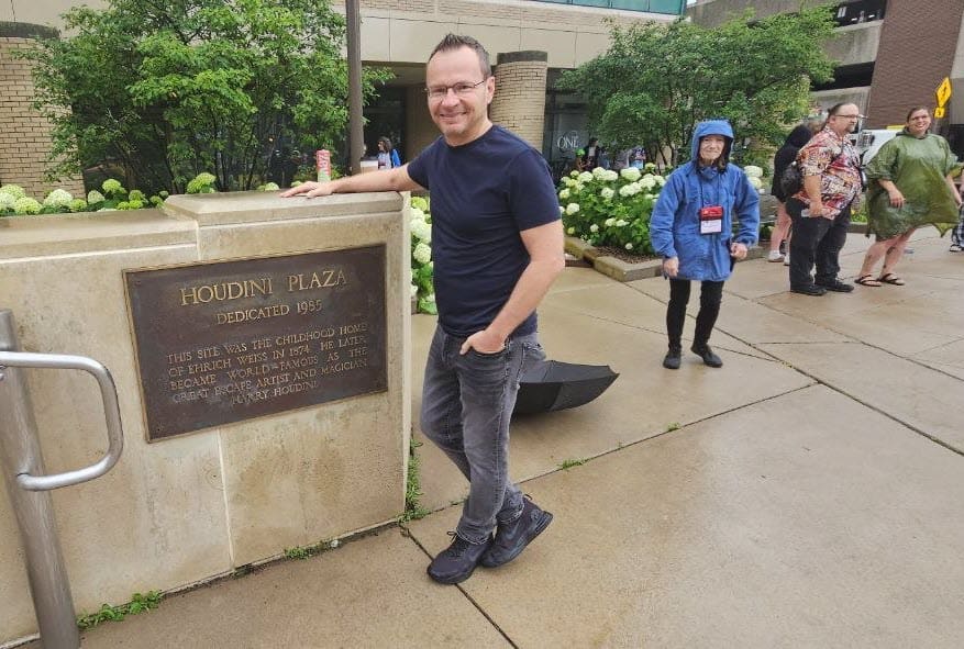 David Farr standing beside the Houdini Plaza dedication marker in downtown Appleton, Wisconsin, umbrella folded at his side